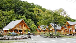A row of wooden cabins with sloped roofs, set against a backdrop of dense green forest. - Cottage Primal Stage (Kamishihoro)