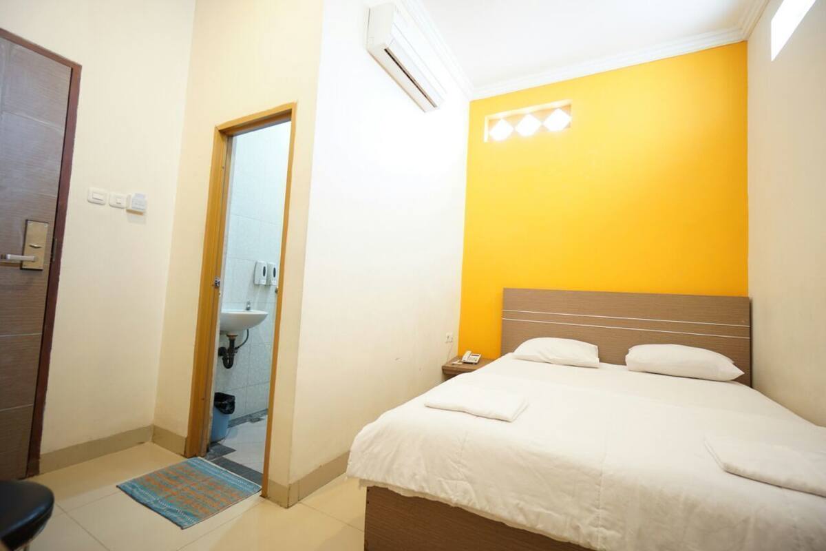Quarto de hotel com cama, mesa de cabeceira, banheiro visível por uma porta aberta e um tapete pequeno no chão.