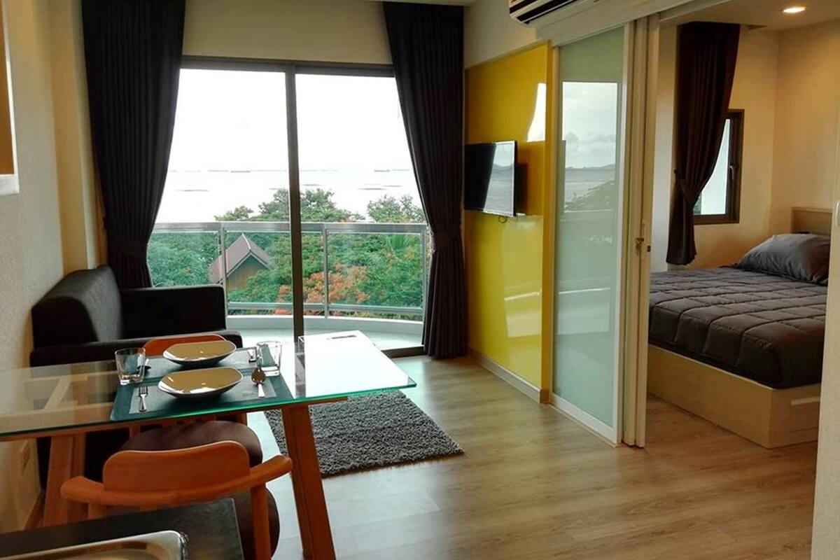Una habitación de hotel moderna con una mesa de comedor de vidrio, una cama individual, un televisor montado en la pared y un balcón con vistas a zonas verdes.
