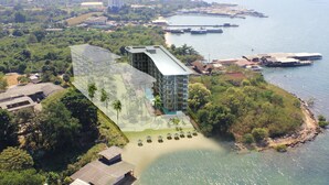 An aerial view of a coastal hotel with a modern design, featuring a large building with a flat roof, surrounded by lush greenery and a beach area with sun loungers. - Sonrisa Sriracha (Si Racha)