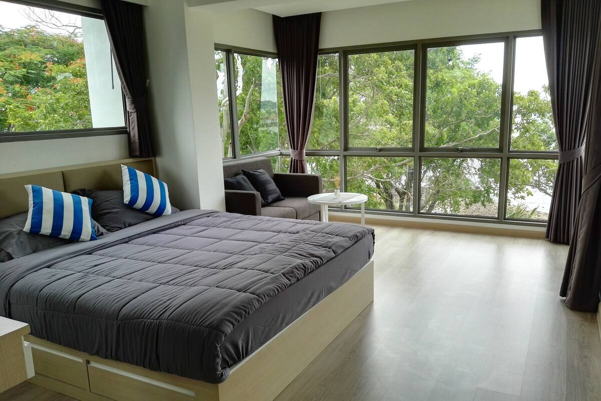 Un dormitorio moderno con una cama grande, un sofá y una mesita. Hay ventanas amplias con cortinas, y se ve un paisaje de árboles desde el exterior.
