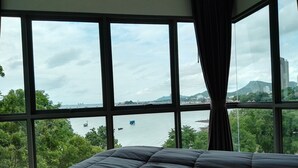 A bed with a dark quilt in a room with large windows overlooking a body of water and distant buildings. - Sonrisa Sriracha (Si Racha)