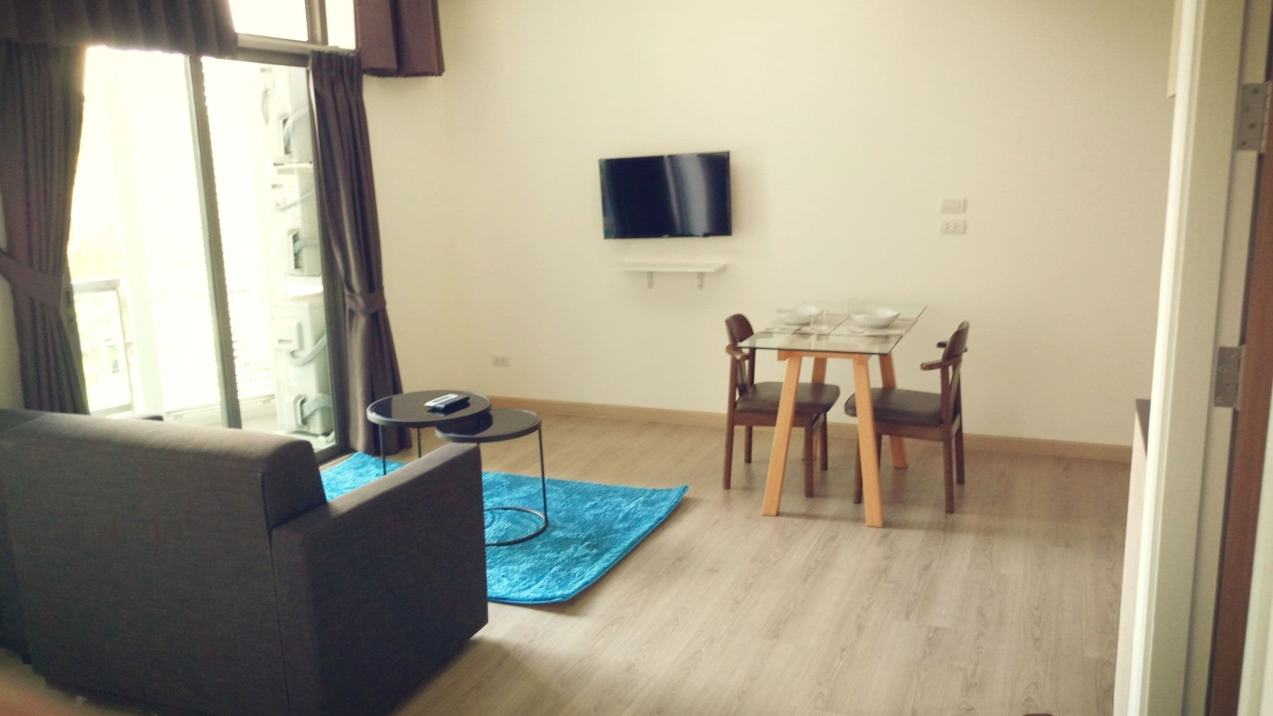 A living room with a sofa, a small round table, a glass dining table with four chairs, a flat-screen TV mounted on the wall, and a blue rug on the floor.