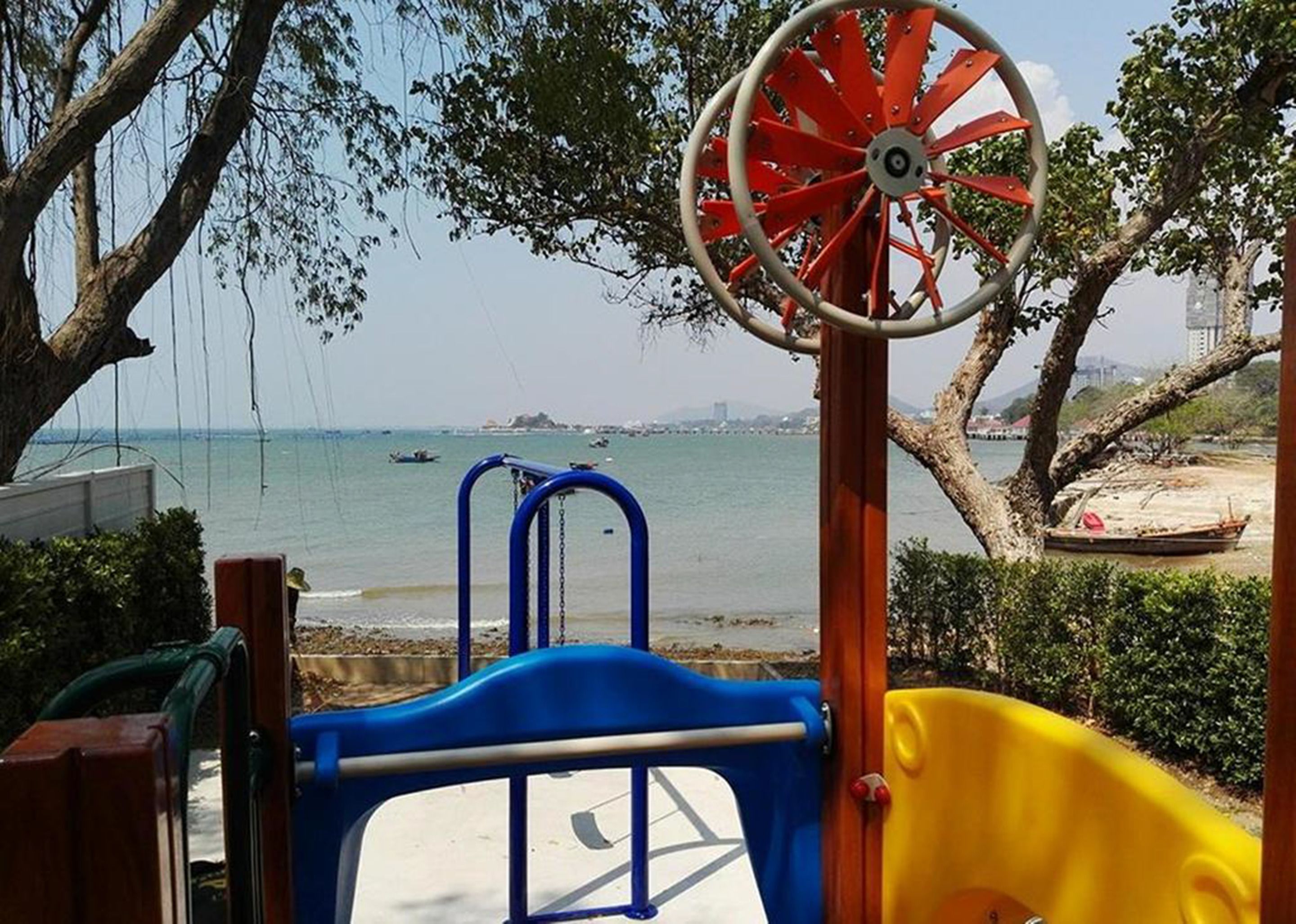 A playground with a slide and a swing set, overlooking a beach with boats and a distant city skyline.