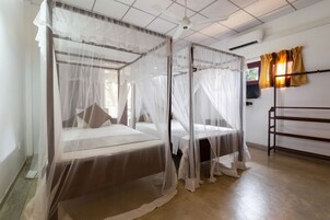 Habitación con dos camas, cada una cubierta por un dosel blanco. Se observa un ventilador de techo, un equipo de aire acondicionado y una ventana con cortinas.