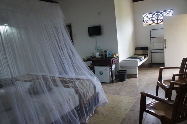 Una habitación con una cama cubierta por una mosquitera, un refrigerador pequeño, un televisor montado en la pared y una ventana con rejas decorativas de hierro.