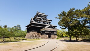 A traditional Japanese castle with multiple tiers and a stone base, surrounded by a paved pathway and trees. - Yukeikohan Suitenkaku (Matsue)