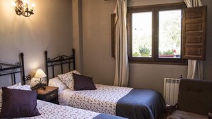 A bedroom with two beds, a nightstand with a lamp, a chair, and a window with curtains. - Casa Rural Los Garridos (Casas Bajas)