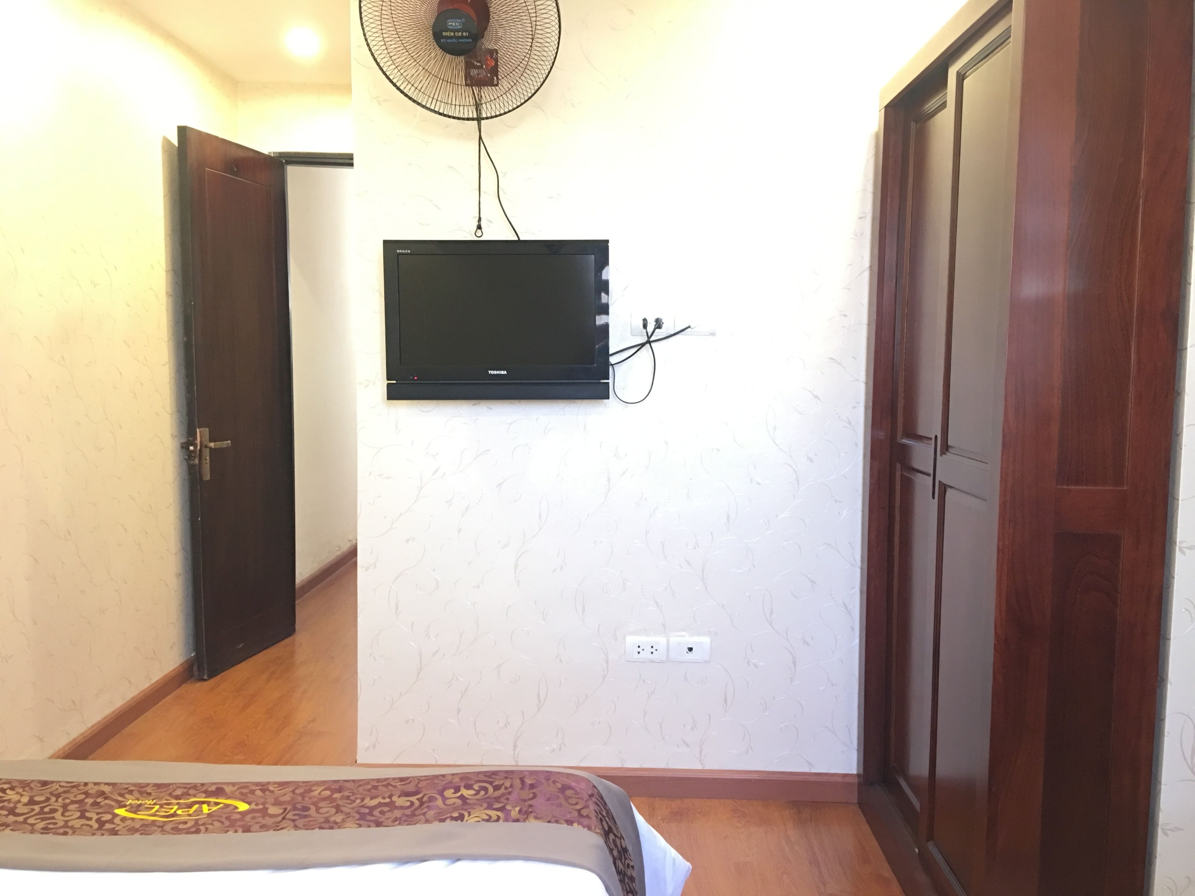 A bedroom with a bed, a wall-mounted TV, a standing fan, and a wardrobe.