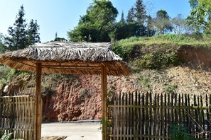 A rustic wooden structure with a thatched roof, surrounded by a bamboo fence and a dirt path leading into a natural landscape.