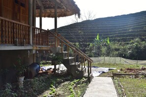 A wooden house with a thatched roof, a staircase leading to a balcony, and a garden with plants and a large drum. - Thong Nguyen Homestay - Hostel (Ha Giang 1)