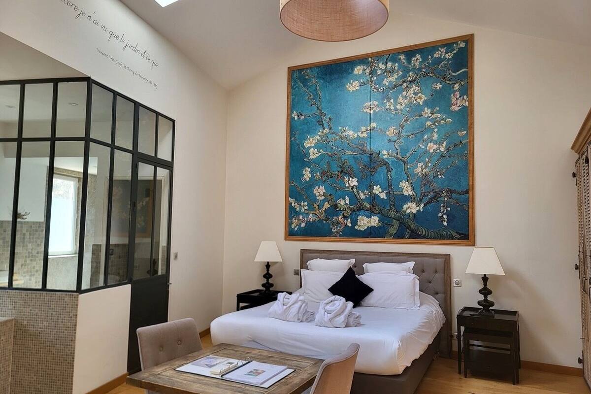 A hotel room with a large bed, two bedside tables, a wooden coffee table, a chair, a large window, and a painting on the wall.