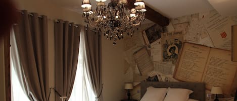 A bedroom with a large bed, a chandelier, a small table with chairs, and a wall covered with papers.
