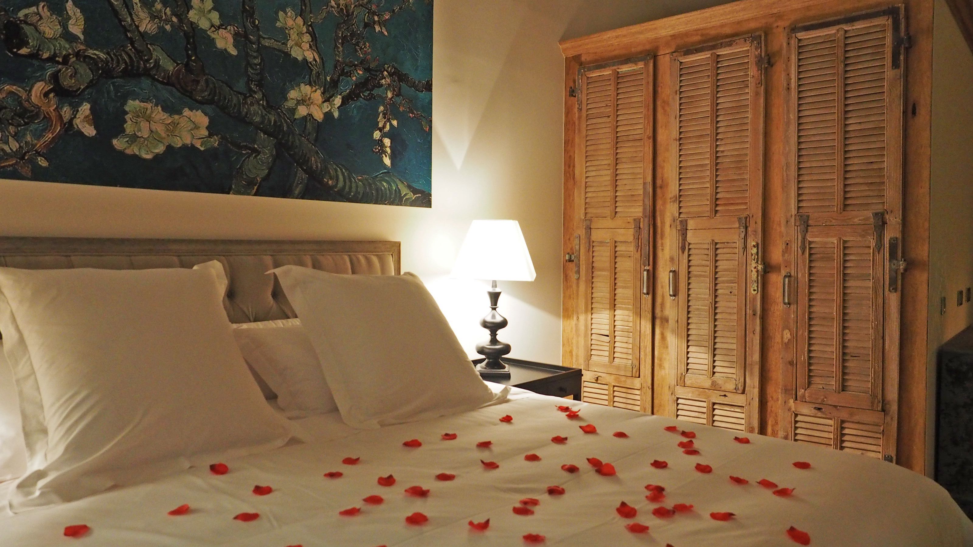 Un lit bien fait, avec des draps blancs et des pétales de roses rouges éparpillés dessus, une lampe sur une table de chevet et un tableau au-dessus du lit.