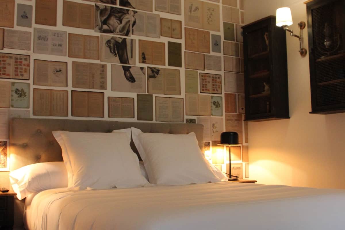 A neatly made bed in a room with a wall covered in framed papers.