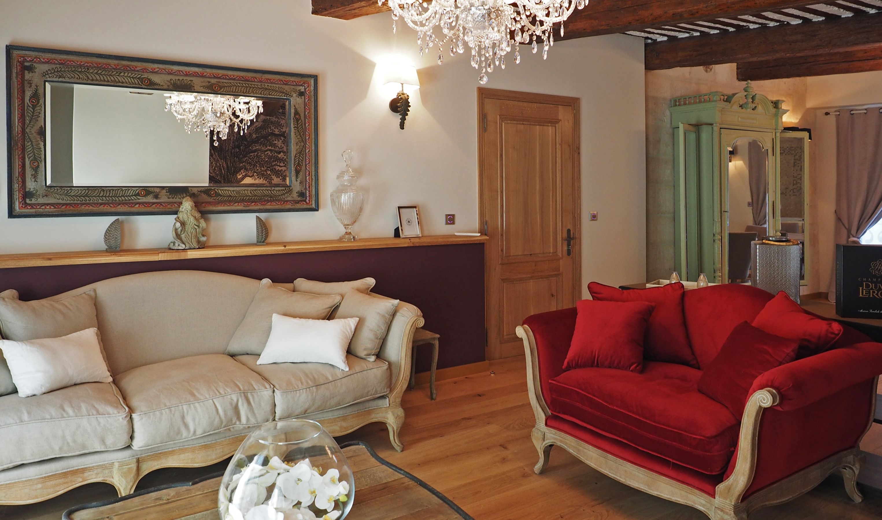 Un salon avec un canapé beige, un canapé en velours rouge, un lustre, un miroir et une porte en bois.