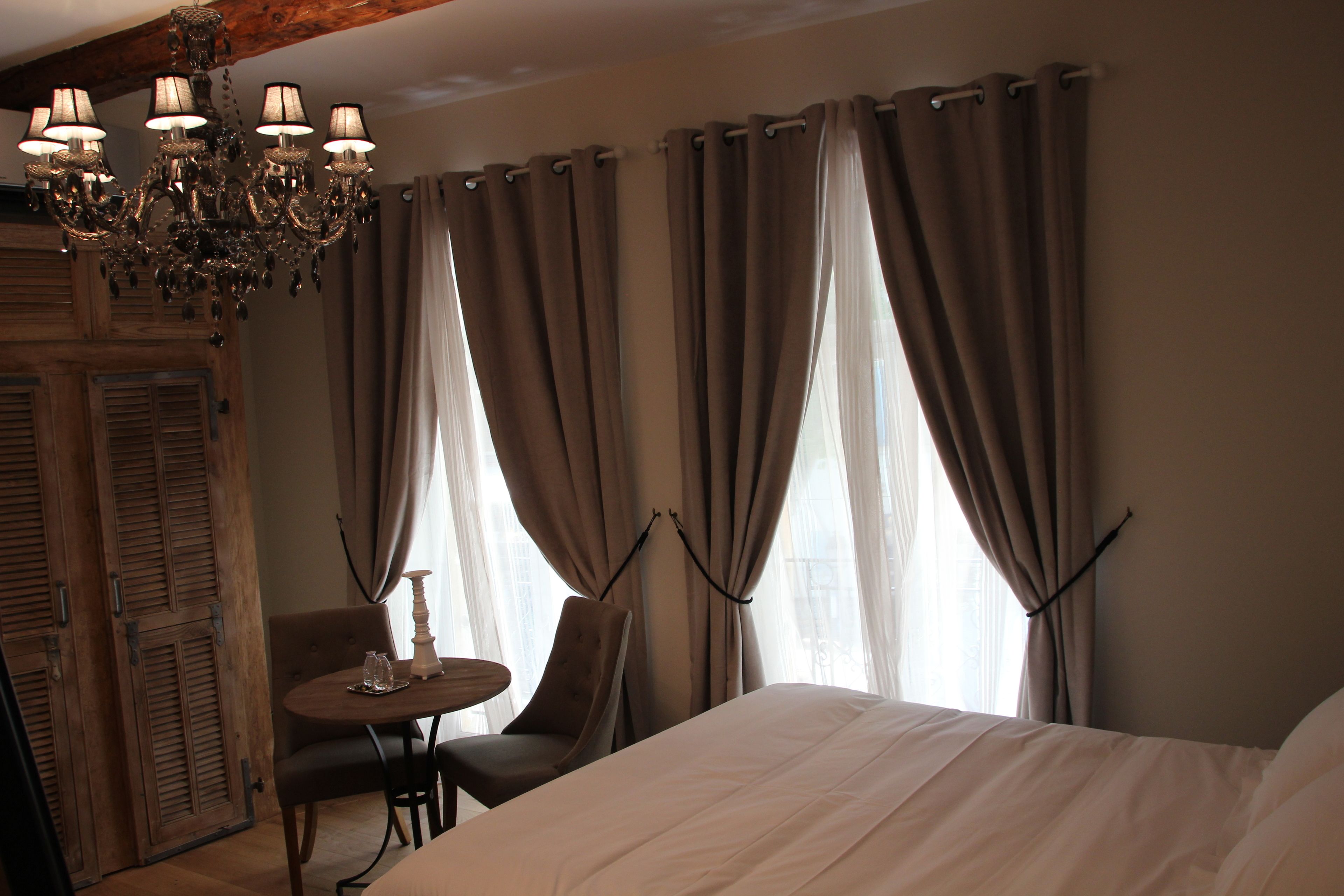 A hotel room with a large bed, a wooden wardrobe, a chandelier, a round table with chairs, and a window with curtains.