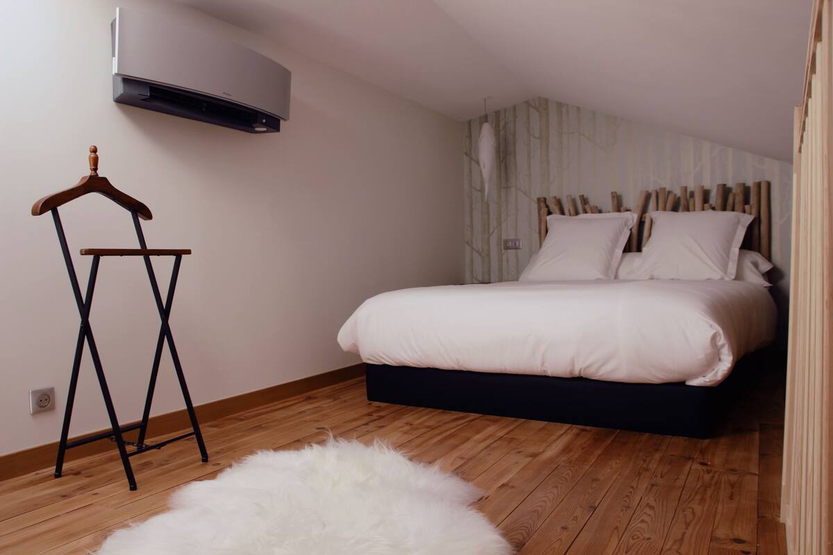 A bedroom with a bed, a wooden floor, a white fluffy rug, a wooden hanger, and a wall-mounted air conditioner.