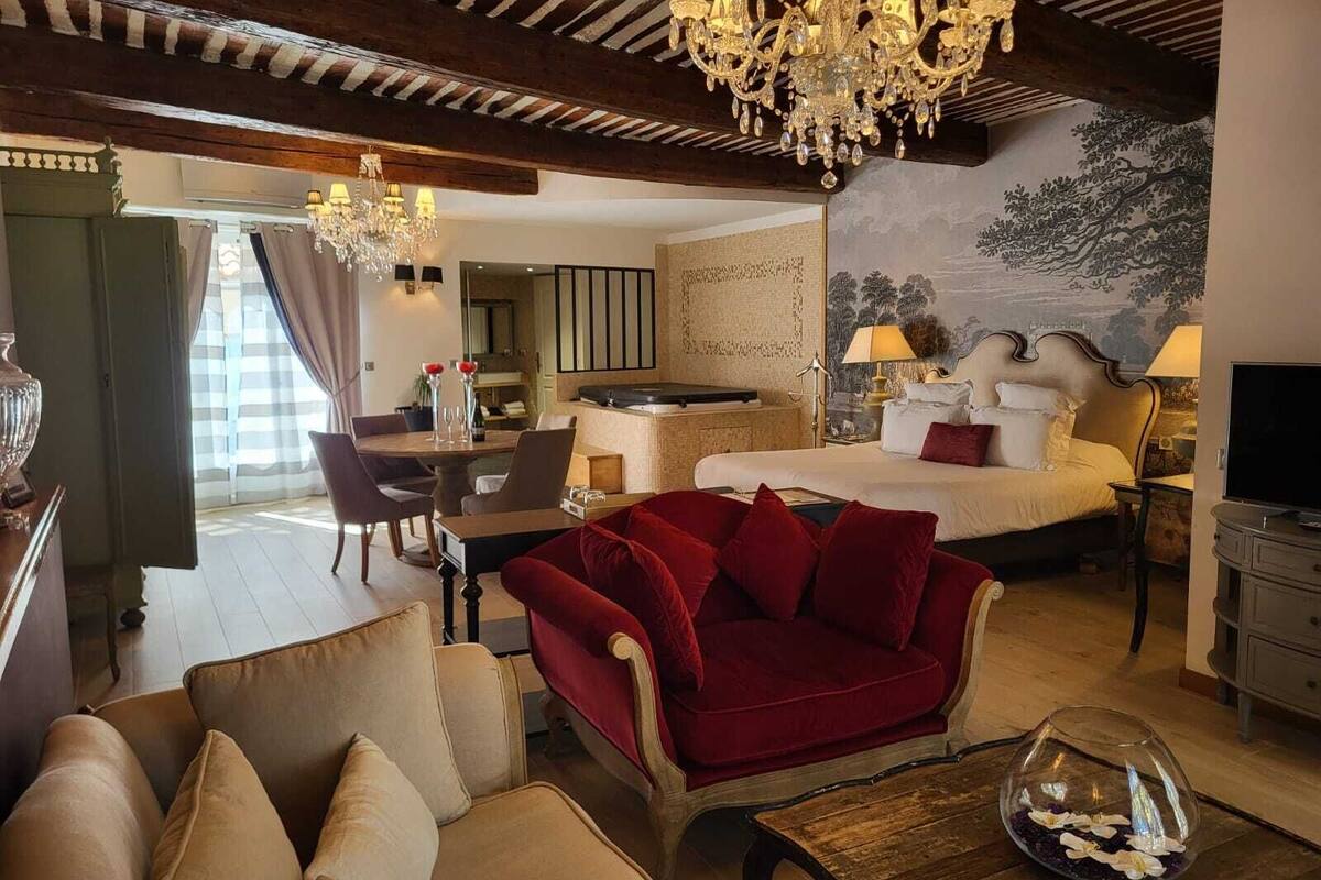 A hotel room with a bed, a red sofa, a dining table with chairs, a chandelier, and a painting on the wall.