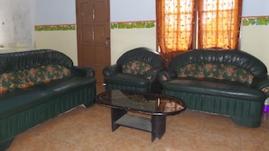 A living room with two green sofas, a glass coffee table, a wooden door, and a window with patterned curtains. - Losmen Setia Kawan II (Ngadisari)