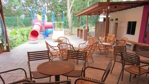 An outdoor patio area with wicker tables and chairs, a playground in the background, and a covered seating area. - Merlin Hotel (Port Dickson)