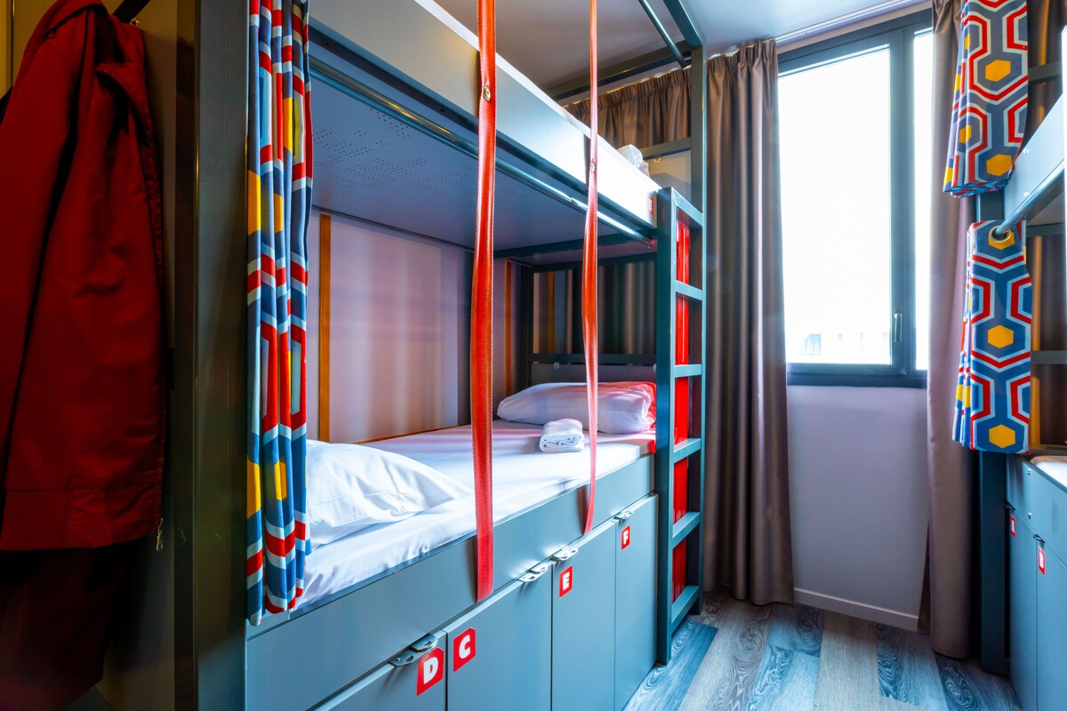 Habitación con literas, una chaqueta roja colgada en la pared, una ventana con cortinas y un armario azul.
