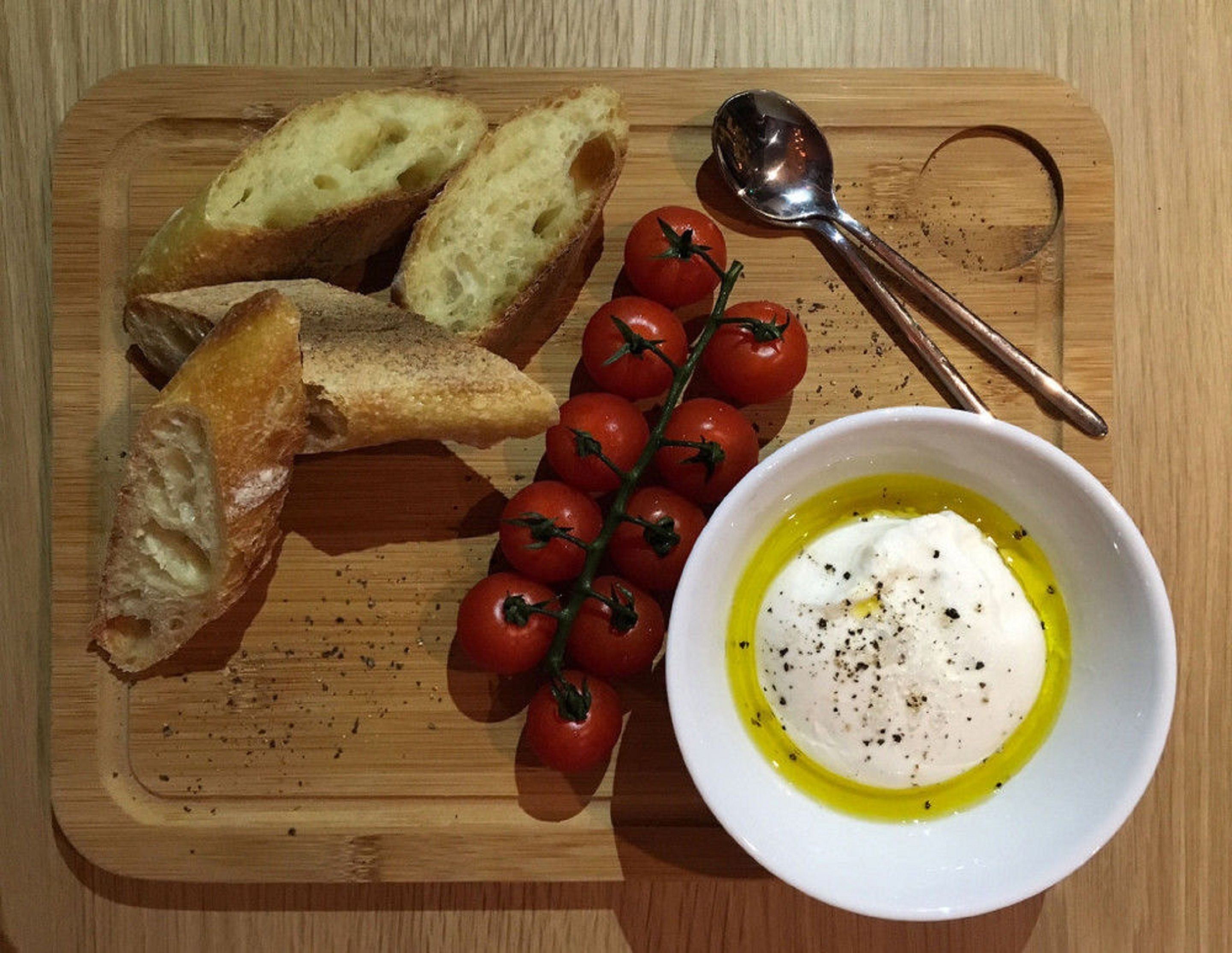 Una tabla de madera con pan en rodajas, tomates cherry y un bol de aceite de oliva y salsa blanca.