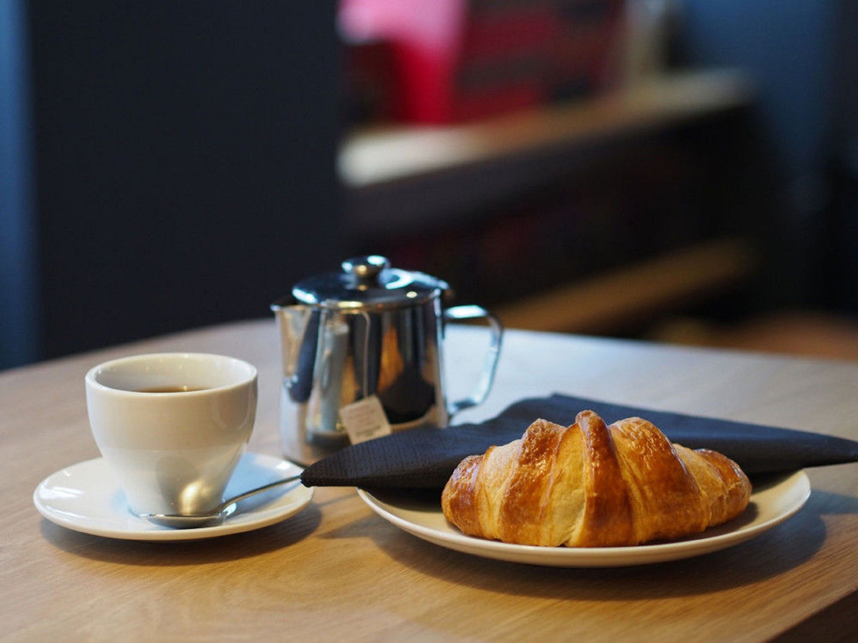 Uma chávena de café num pires, um bule de café de metal e um croissant num prato sobre uma mesa de madeira.