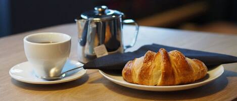 A cup of coffee on a saucer, a metal coffee pot, and a croissant on a plate on a wooden table.
