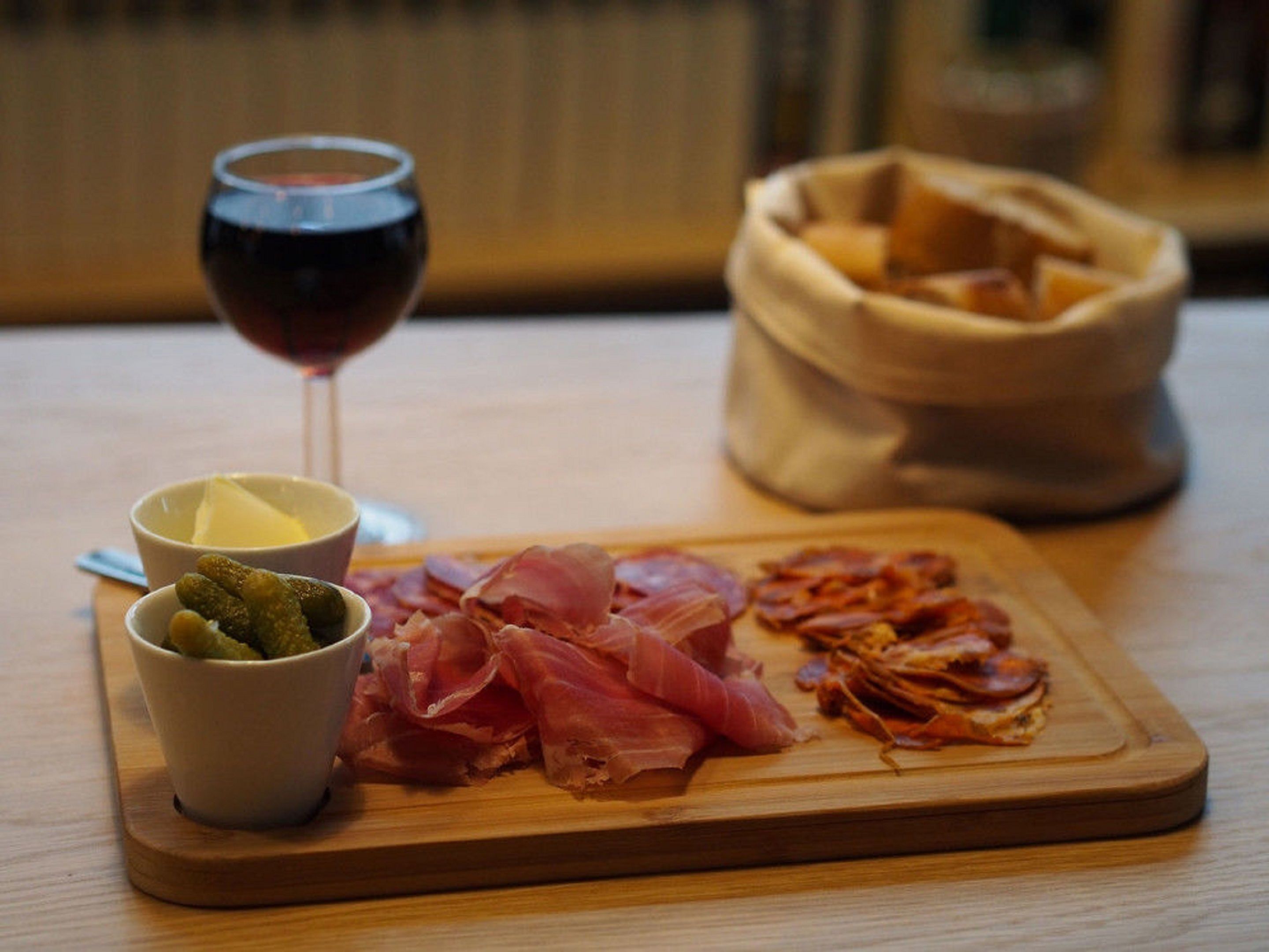 A wooden table with a glass of red wine, a bowl of olives, a small bowl of butter, and a wooden board with sliced cured meats and pickles.
