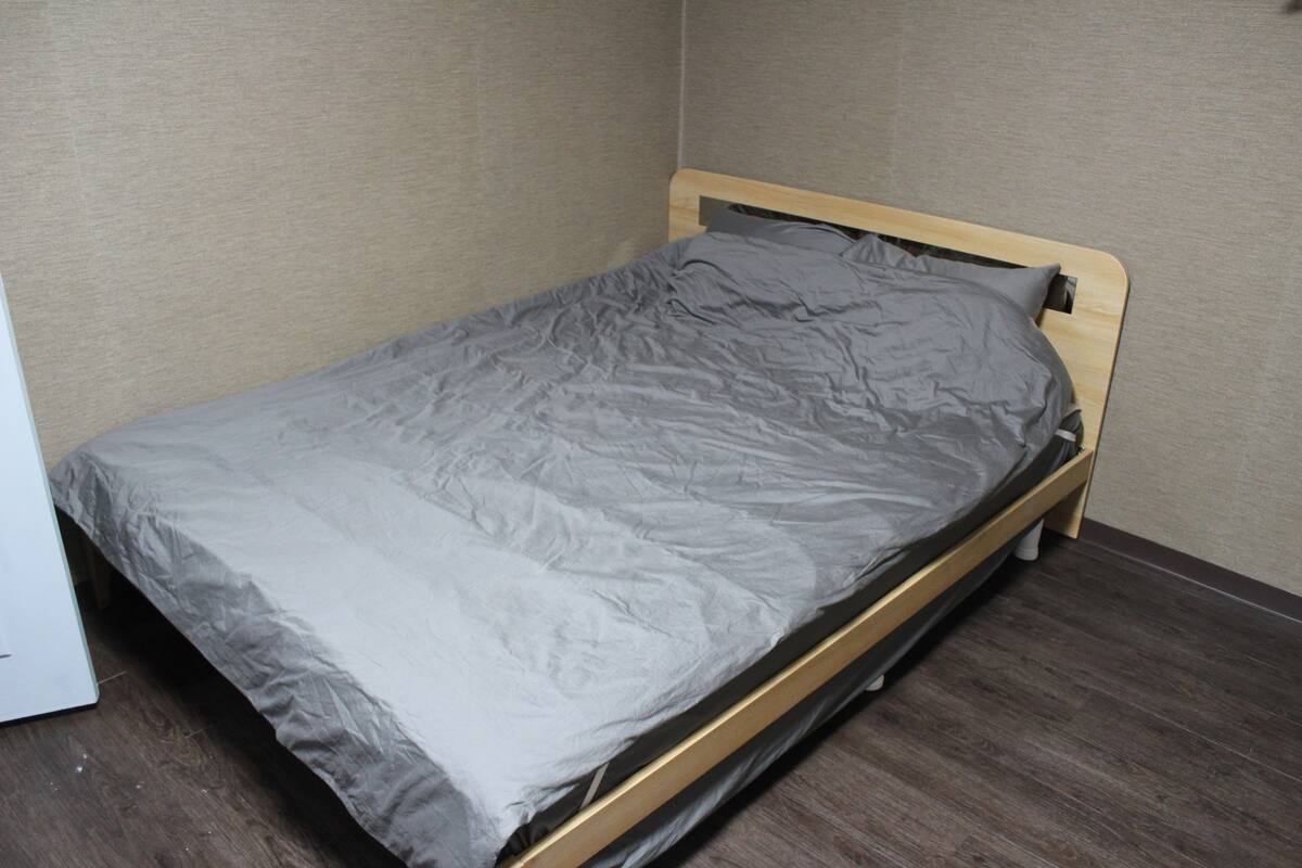 A single bed with a wooden frame and a grey mattress in a room with wooden flooring and beige walls.