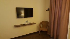 A room with a flat-screen TV mounted on the wall, a wooden shelf with toiletries, a wicker chair, and a brown curtain. - 9 Square Hotel - Kota Damansara (Petaling Jaya)