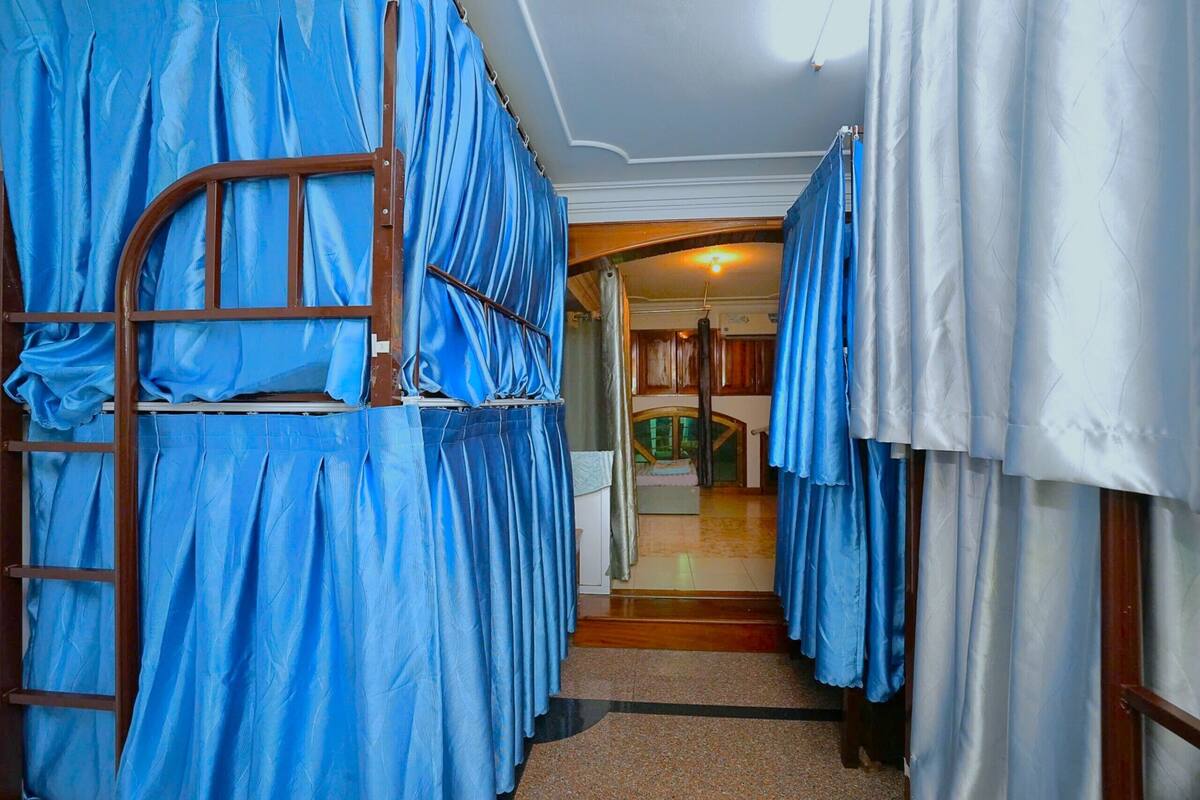 A corridor with bunk beds covered in blue fabric, wooden flooring, and a door leading to a room with a bed and curtains.
