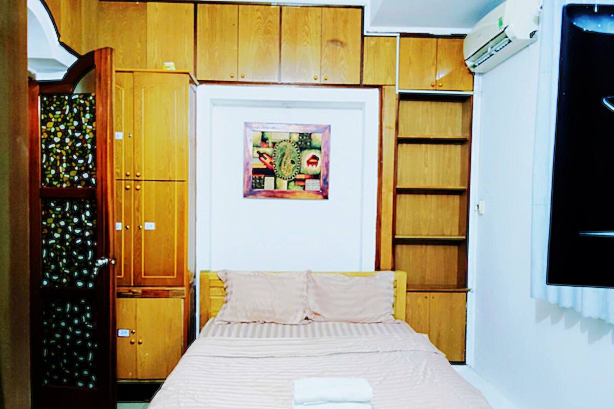 A bedroom with a bed, a wooden wardrobe, a small cabinet, and a wall-mounted air conditioner.