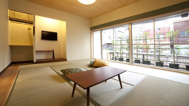 A traditional Japanese room with tatami flooring, a low wooden table, a television on the wall, and a view of the street through large windows. - Fukuwauchi (Tokyo)