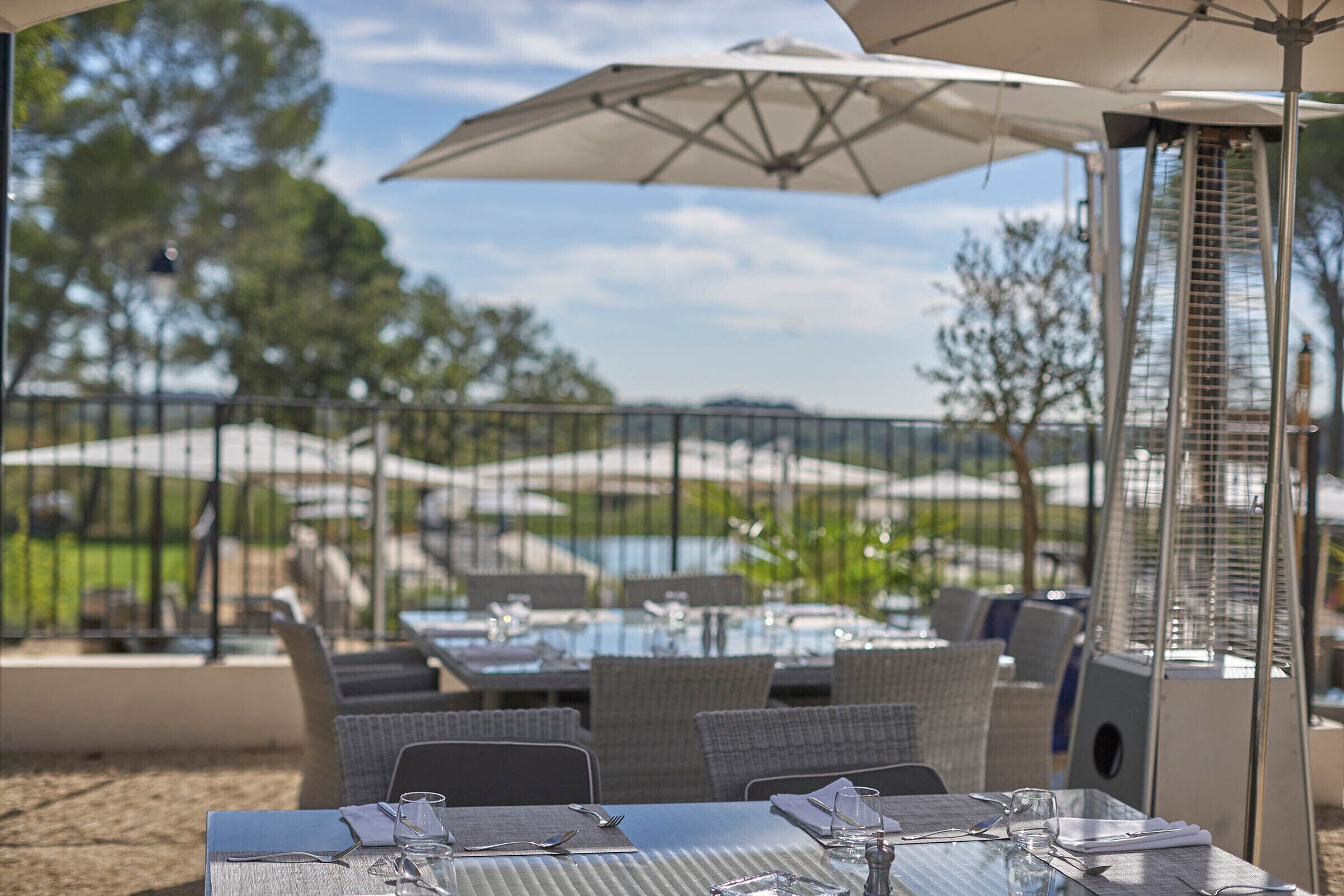 Un espacio de comedor al aire libre con mesas y sillas, un paraguas grande y vistas a un campo de golf.