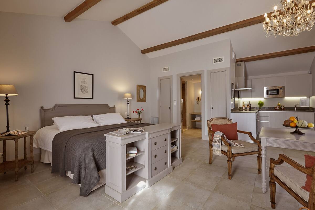 A bedroom with a bed, bedside tables, a desk, and a chair. There is a kitchen area with a dining table and chairs.
