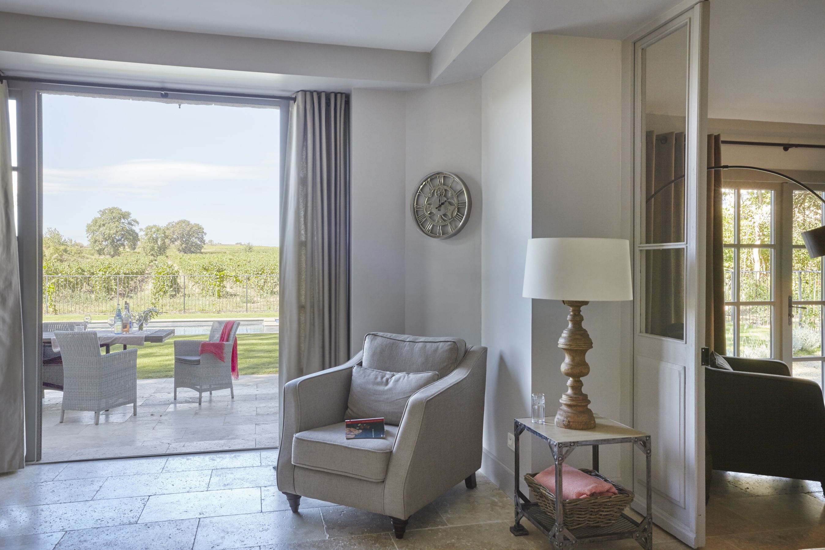 A living room with a grey armchair, a table with a lamp, and a view of a garden through a sliding door.