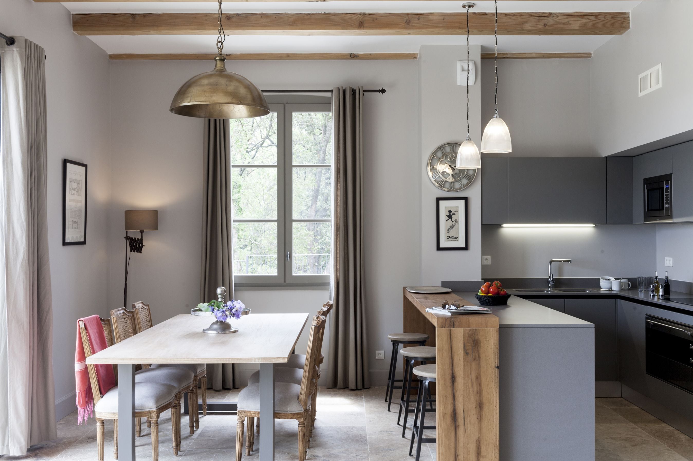 A modern kitchen with a central island, wooden beams, pendant lighting, and a large window with curtains.
