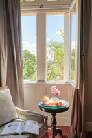 Una habitación con vistas a la vegetación por la ventana, una mesa con una bandeja de comida y bebidas, y un libro en una silla.