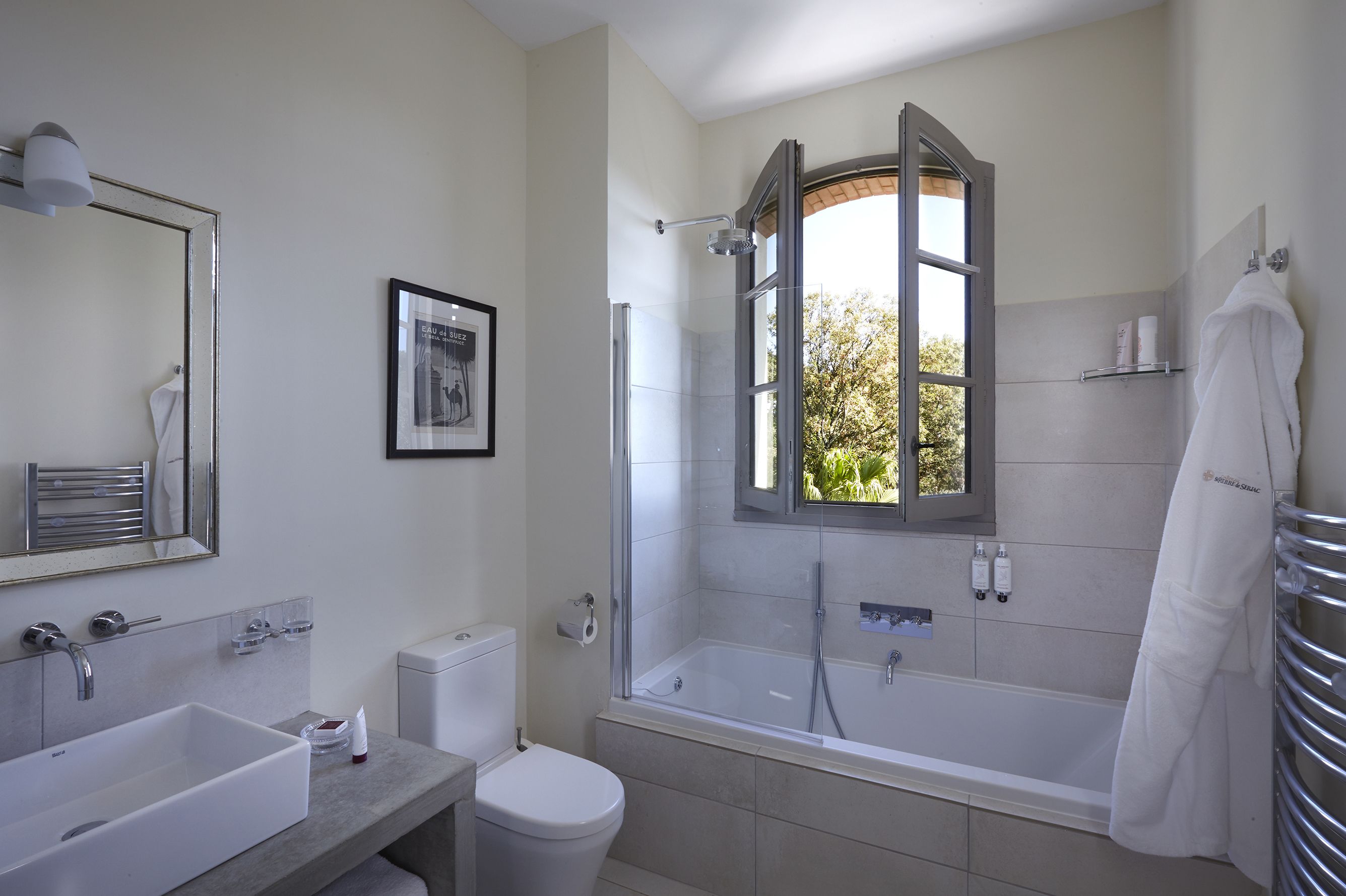 A modern bathroom with a large bathtub, a toilet, and a sink. There's a window with an open door, a towel rack, and a framed picture on the wall.