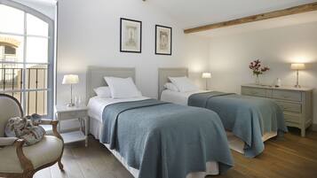 Habitación de hotel con dos camas, una silla de madera, una mesita de noche con lámpara y un aparador con un florero.