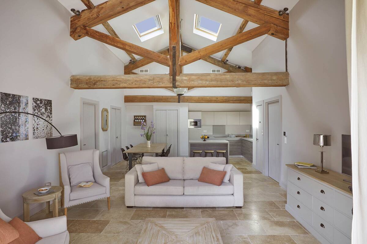 A spacious living area with a high ceiling and exposed wooden beams, a white sofa with two cushions, a small side table, and a dining area in the background.