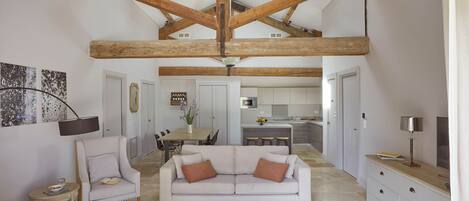 A spacious living area with a high ceiling and exposed wooden beams, a white sofa with two cushions, a small side table, and a dining area in the background.