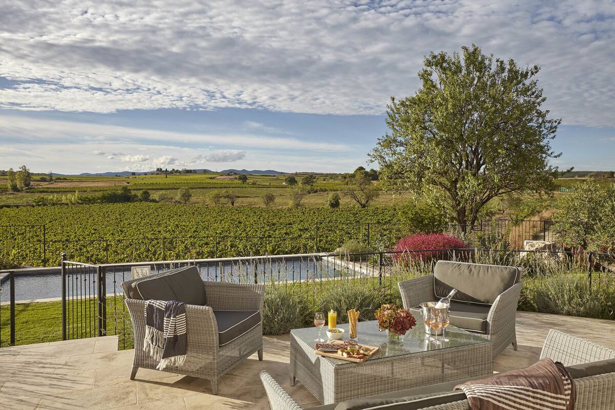 An outdoor patio with wicker furniture, a glass table, and a view of a vineyard and river.