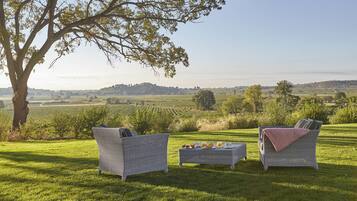 Un espacio exterior con césped, con dos sillas de mimbre y una mesa dispuesta para una comida, con vistas a un paisaje rural.