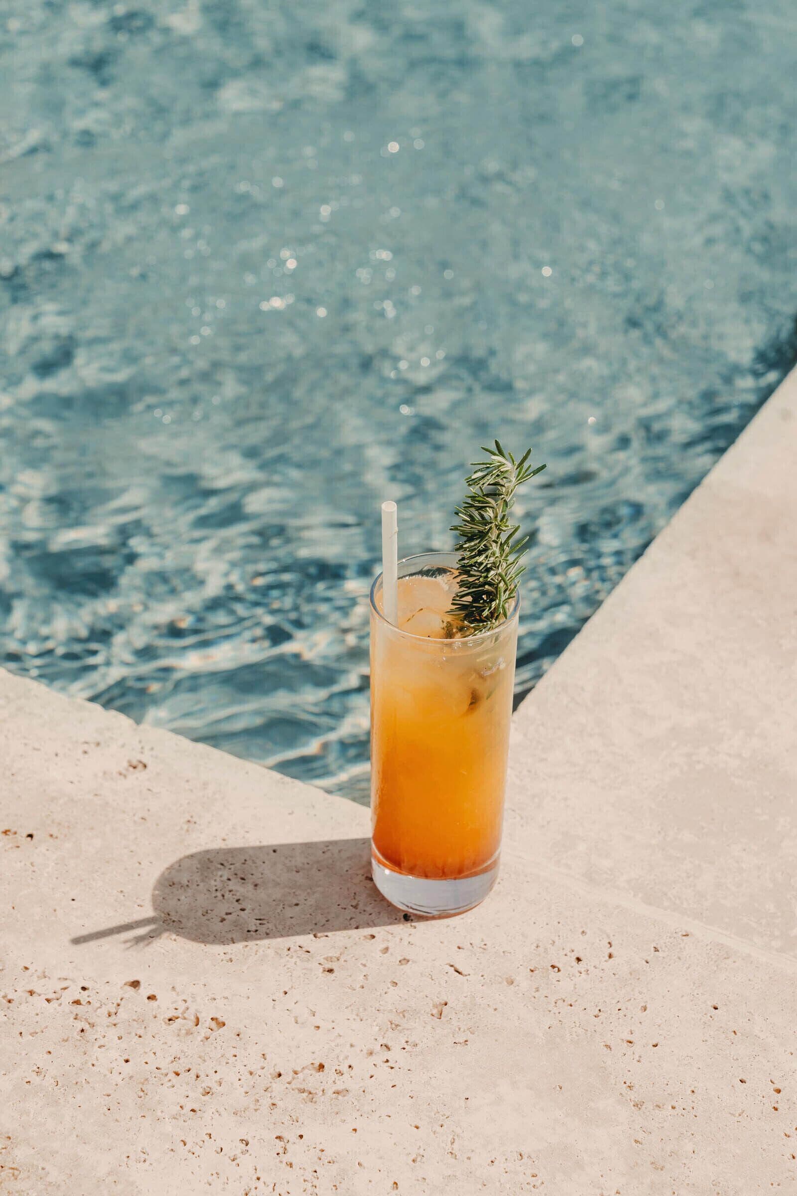A glass of amber-colored drink with a rosemary garnish on a concrete surface by a pool.