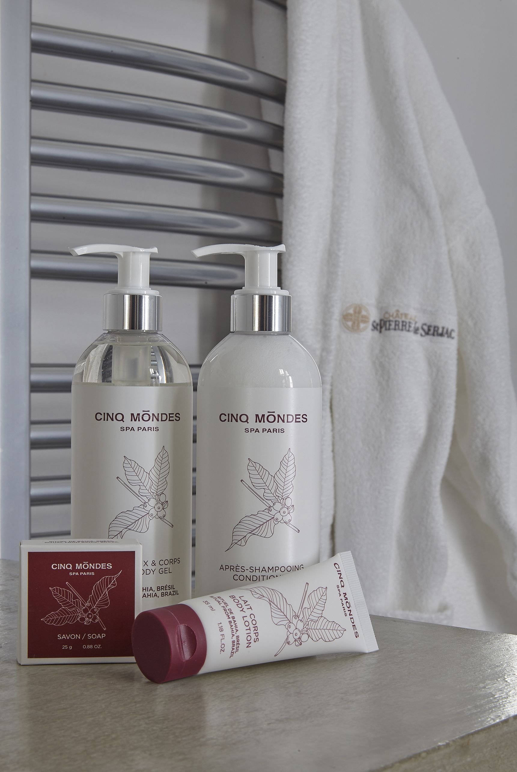 Two bottles of Cinq Mondes spa products, a tube of shampoo, and a red box of soap, all placed on a table with a white towel in the background.