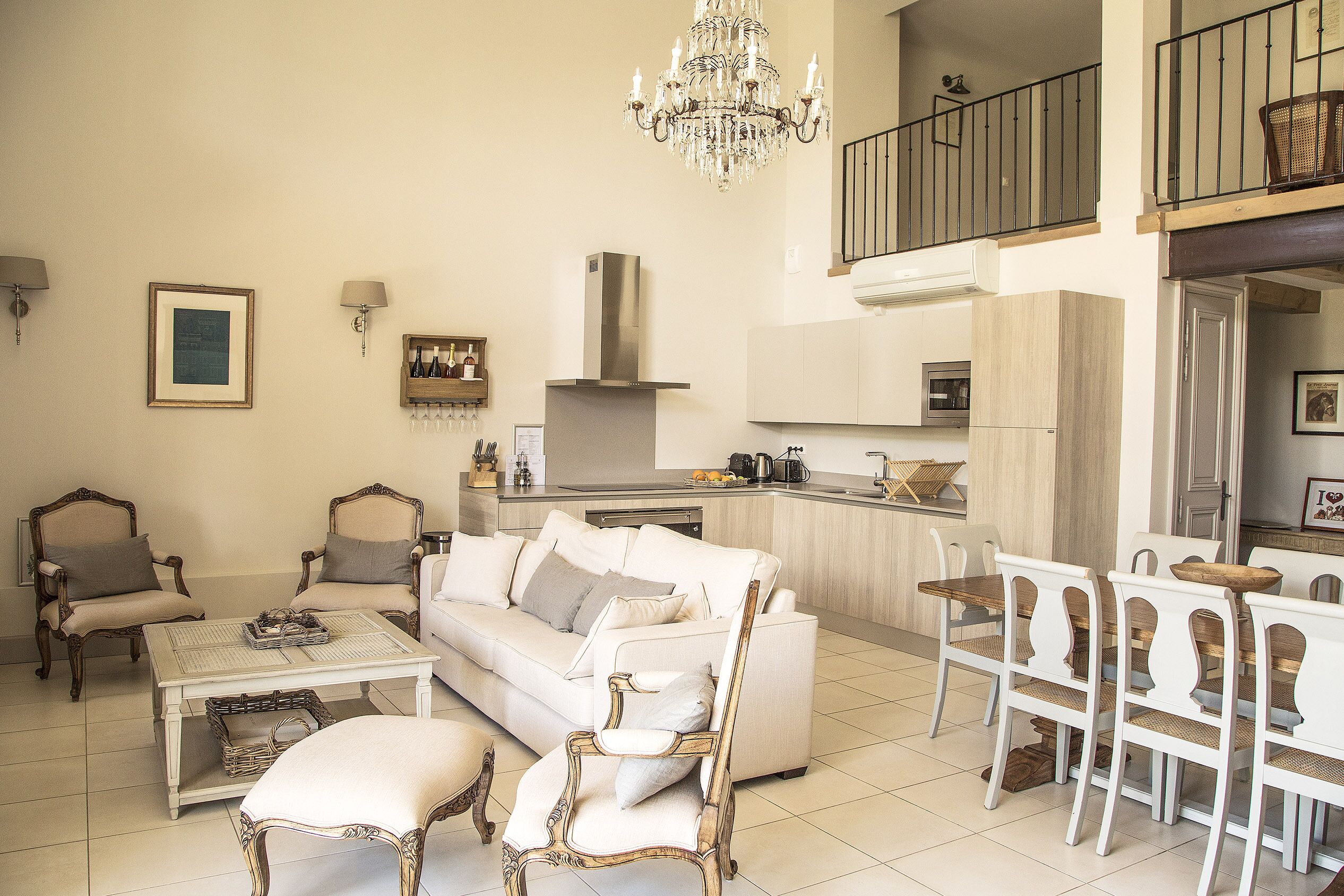 A spacious living room with a white sofa, two armchairs, a dining table with chairs, a chandelier, and a kitchen area in the background.