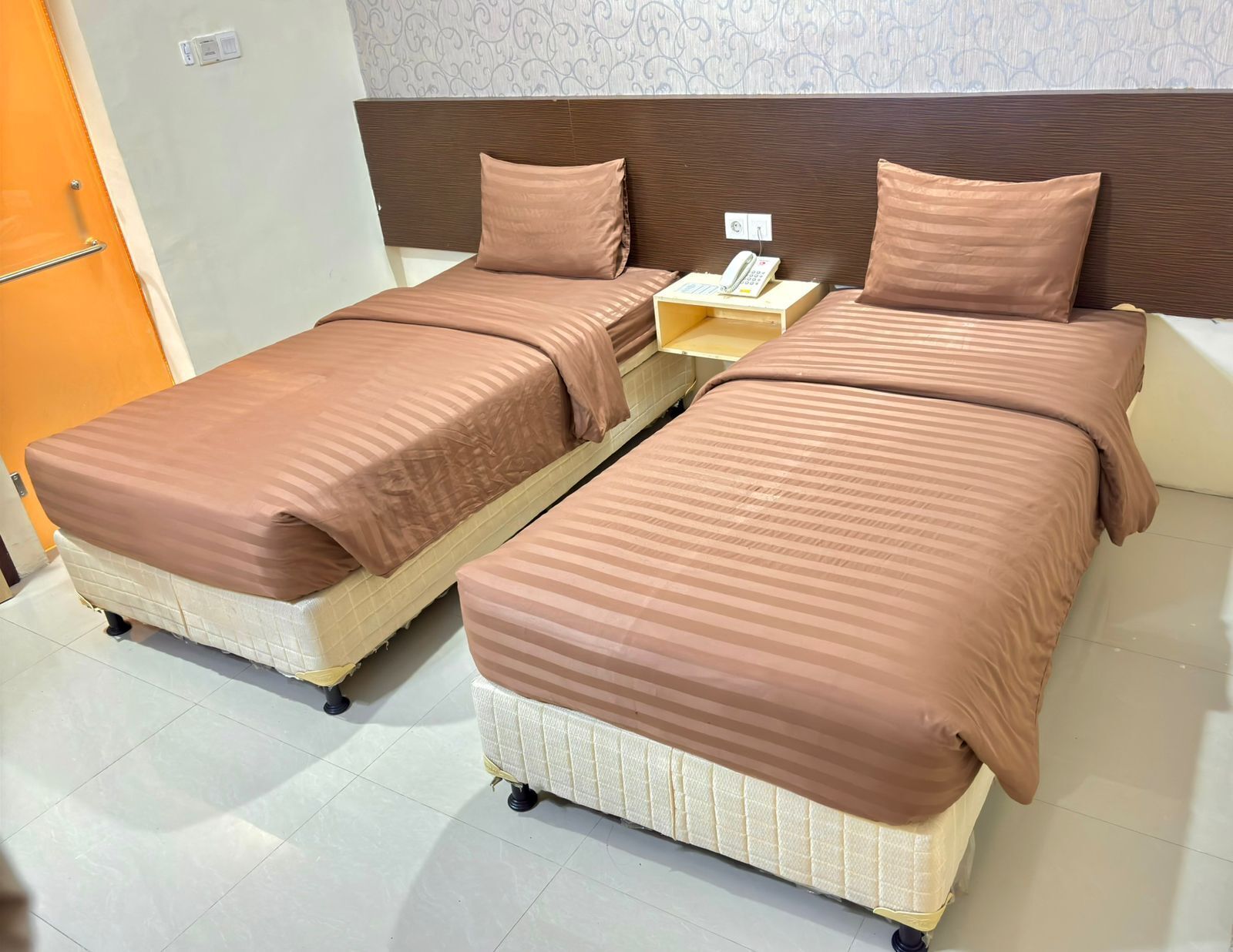 A double room with two beds, each with a headboard, a bedside table with a phone, and a wall-mounted light switch.
